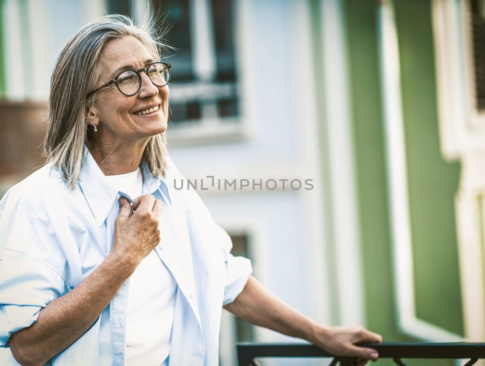 Happiness in old age, mature woman with radiant smile standing on balcony overlooking old street in Europe. Senior lady in joyful and contented state, embracing beauty of aging gracefully. by LipikStockMedia