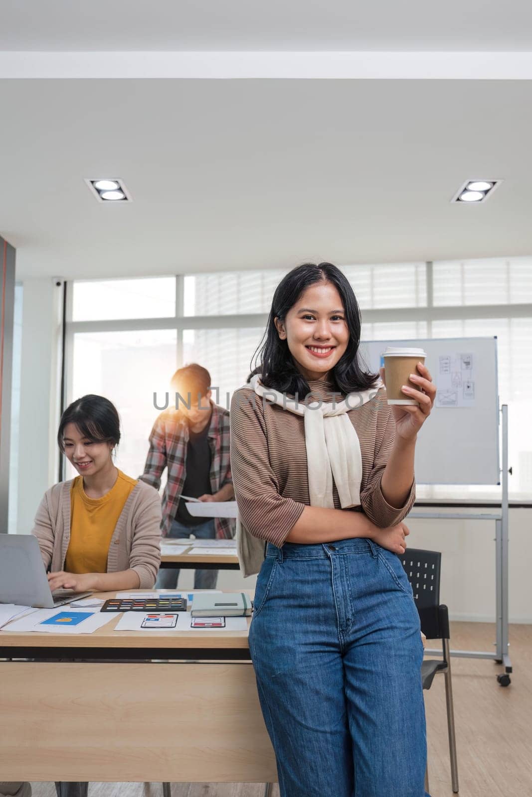 young Asian female marketing assistant is working on her work on her laptop at her desk while her coworkers are working in the room