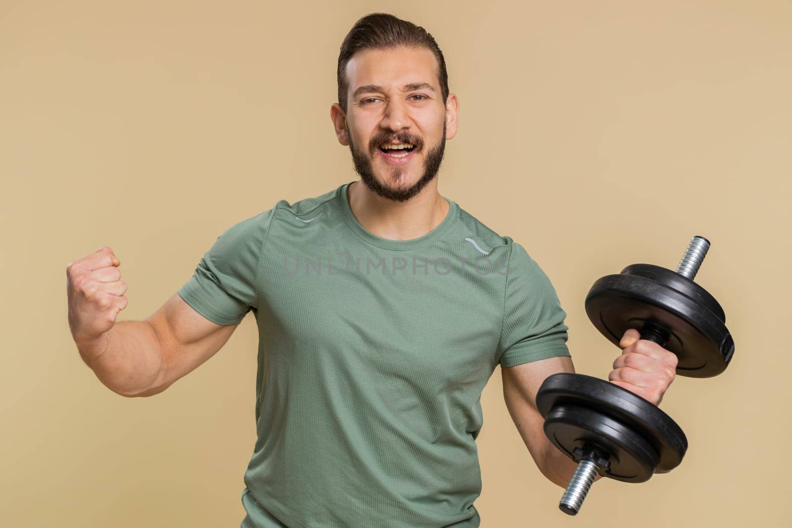 Excited athlete sportive man shouting, celebrating success, winning, goal achievement, good news, lottery luck, wow reaction. Middle eastern sportsman guy weightlifting dumbbell on beige background