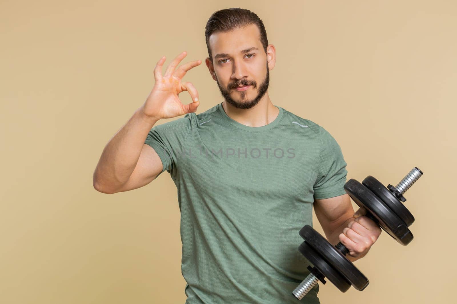 Okay. Athlete sportive man looking at camera showing ok gesture, like sign positive, approve something good, feedback, celebrate, agree. Middle eastern sportsman guy with dumbbell on beige background