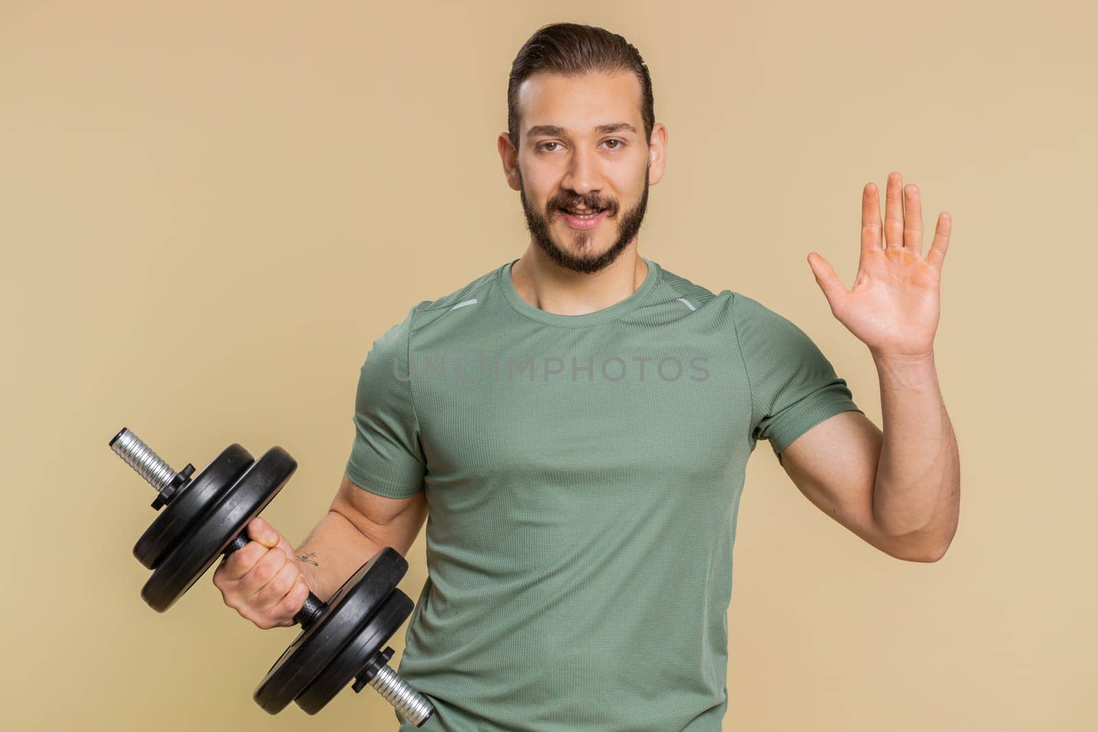Come here, join us. Welcome. Athlete sportive man showing inviting gesture with hands, ask to join, beckoning to coming, gesturing hello or goodbye. Sportsman guy with dumbbell on beige background
