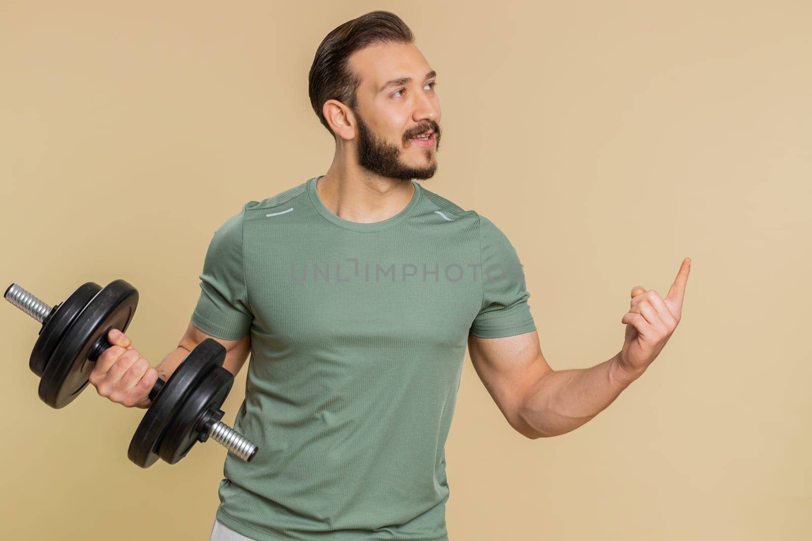 Athlete man showing pointing empty place, advertising area for commercial text, copy space for goods promotion, recommendation. Middle eastern sportsman guy weightlifting dumbbell on beige background