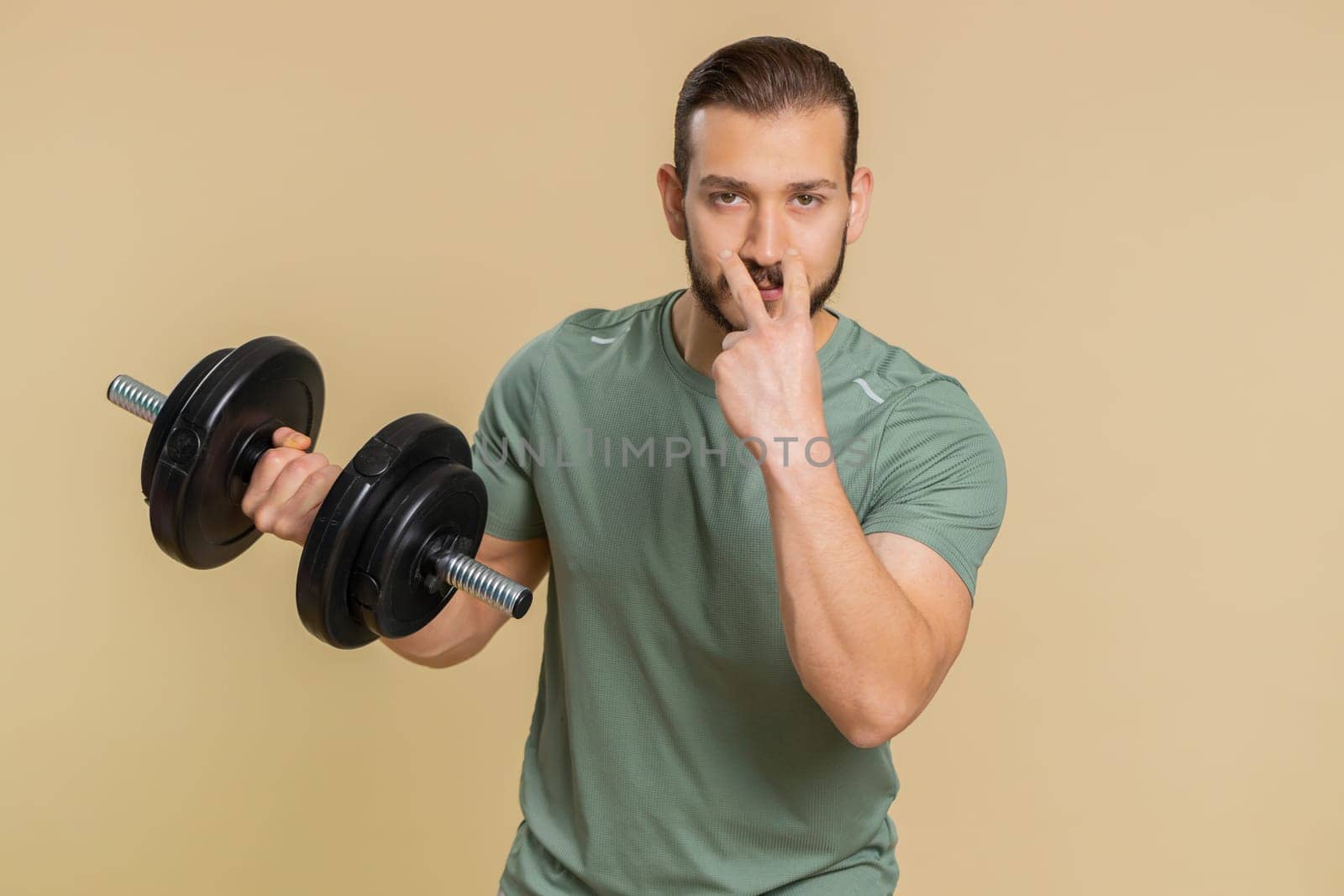 I am watching you. Athlete sportive man pointing at eyes and camera, show I am watching you gesture, spying on someone start working out. Middle eastern sportsman guy with dumbbell on beige background