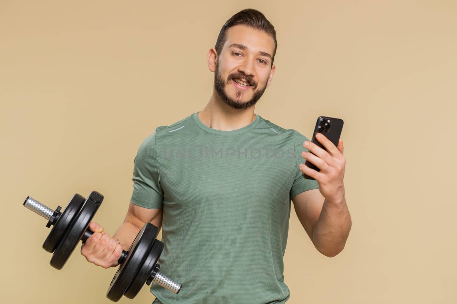 Athlete sportive man texting share messages content on smartphone social media applications online, sms message, browsing, reading news. Middle eastern sportsman guy with dumbbell on beige background