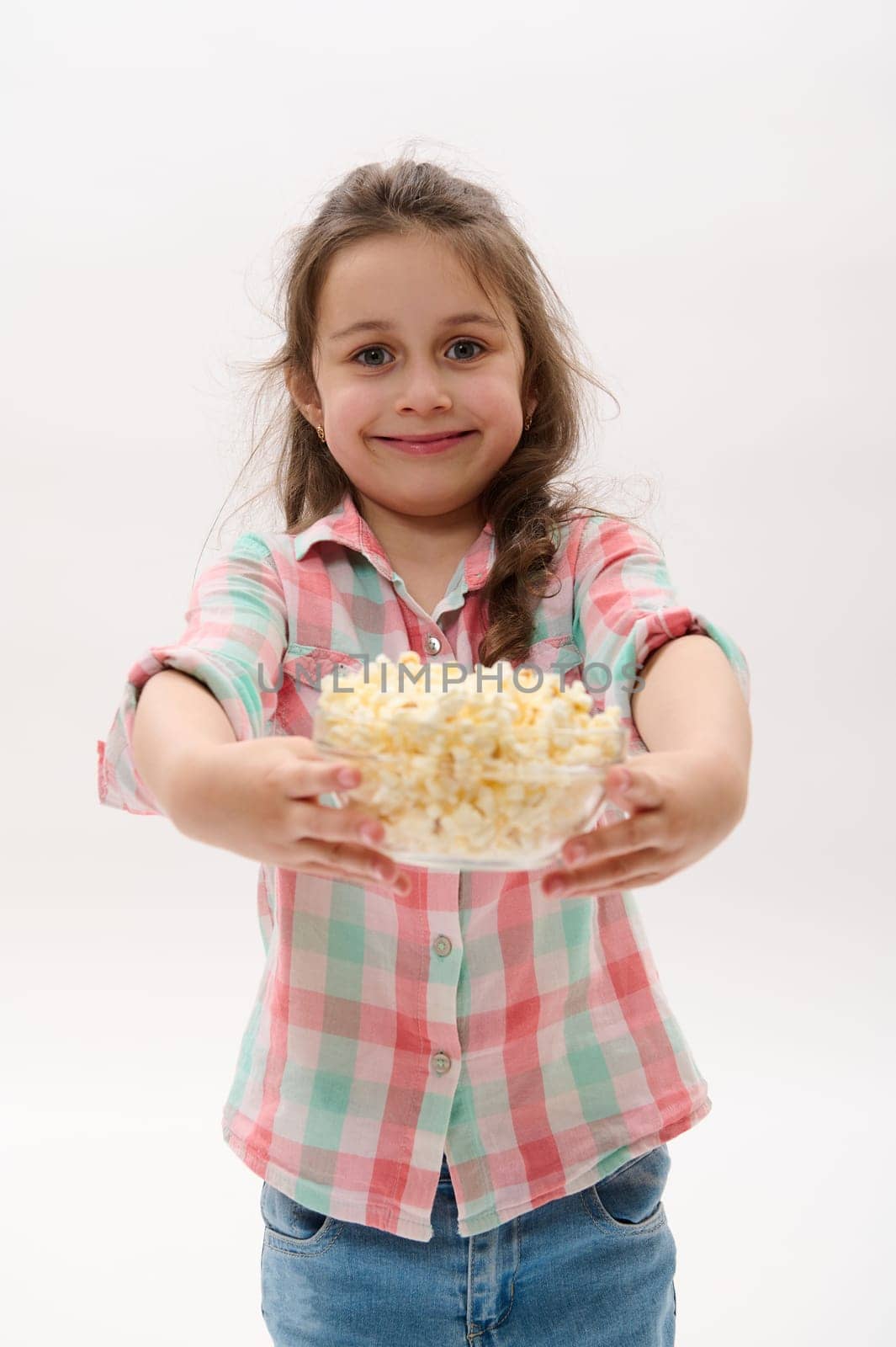 Happy cheery Caucasian little girl in casual clothes, holding out at camera a bowl with salted popcorn, ready to watch cartoon or movie, smiling on white background. People. Kids. Food. Entertainment
