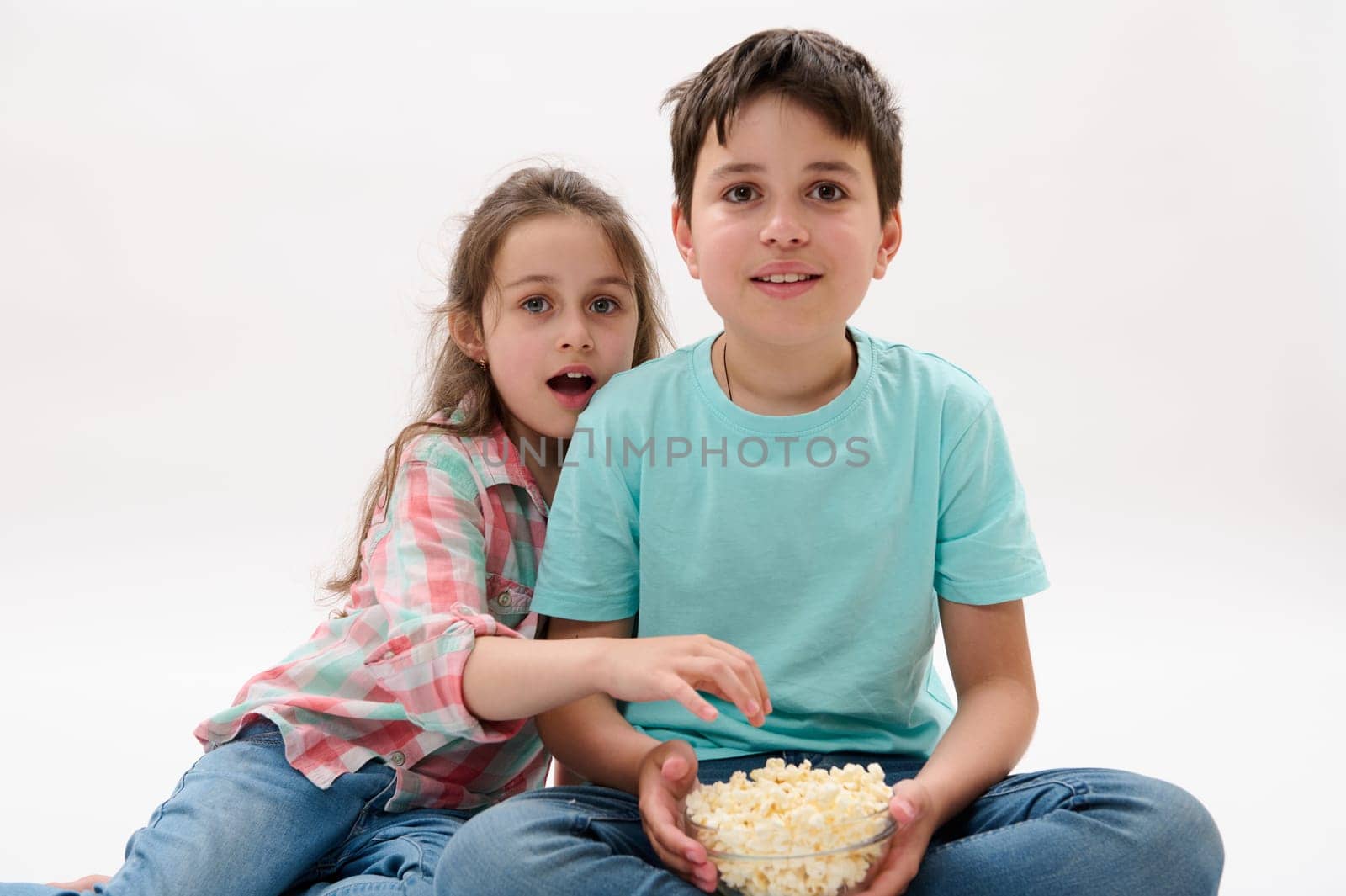 Two amazed kids, teenage boy and little girl, brother and sister watching movie, eating popcorn, expressing wow emotion by artgf