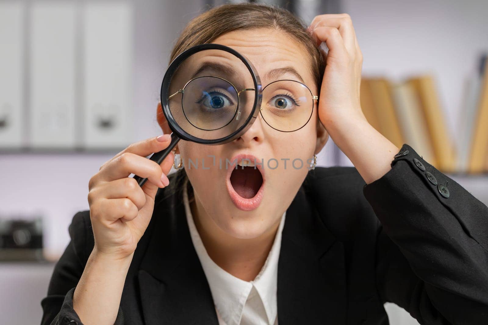 Investigator researcher scientist business woman working at office holding magnifying glass near face looking into camera with big zoomed funny eye, searching, analysing. Professional freelancer girl