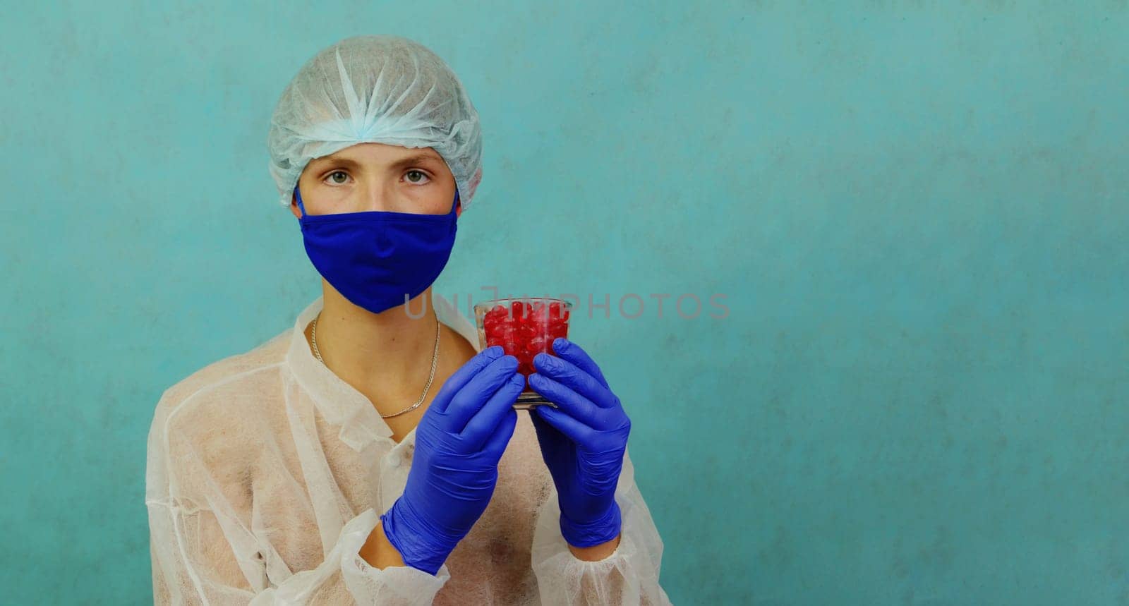 A guy in blue gloves and hat and a white coat is holding a glass of red pills against a studio background. by gelog67