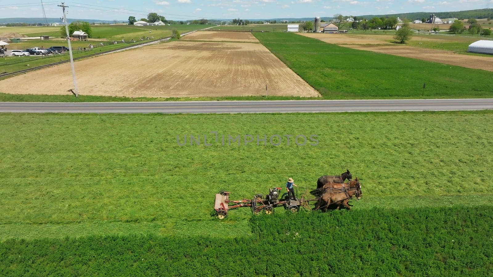 An Aerial View of an Amish Farmer Cutting Mowing Crops Using Four Horses and a Gas Engine With his Barn and Farm House in View, on a Beautiful Summer Day