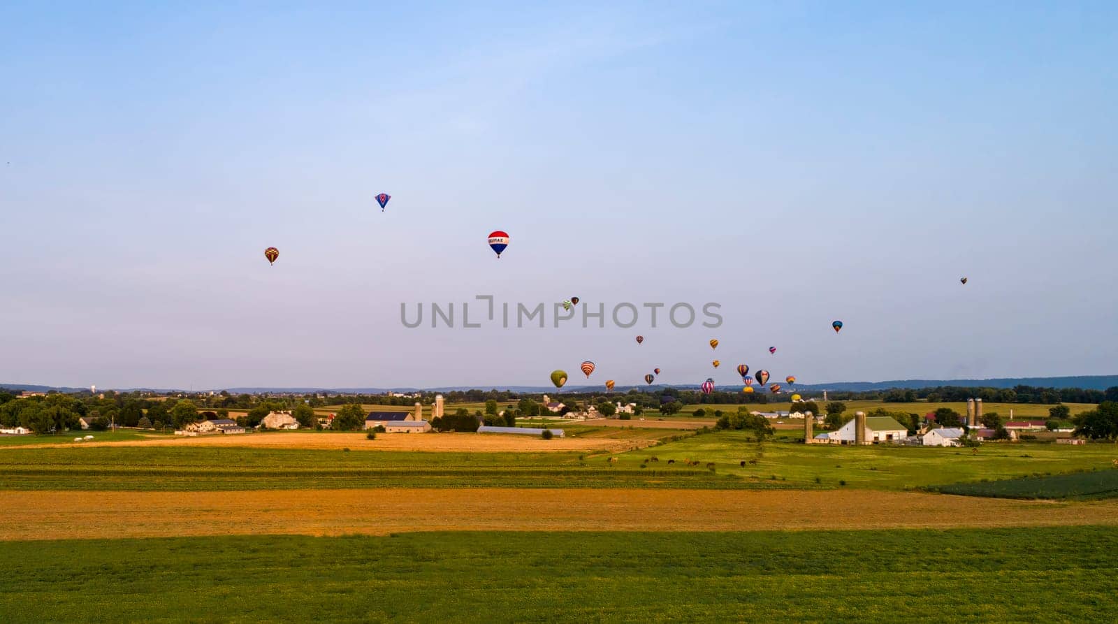 An Drone View of Multiple Colorful Hot Air Balloons Floating Over Farmlands and Countryside on a Beautiful Summer Day, During a Festival