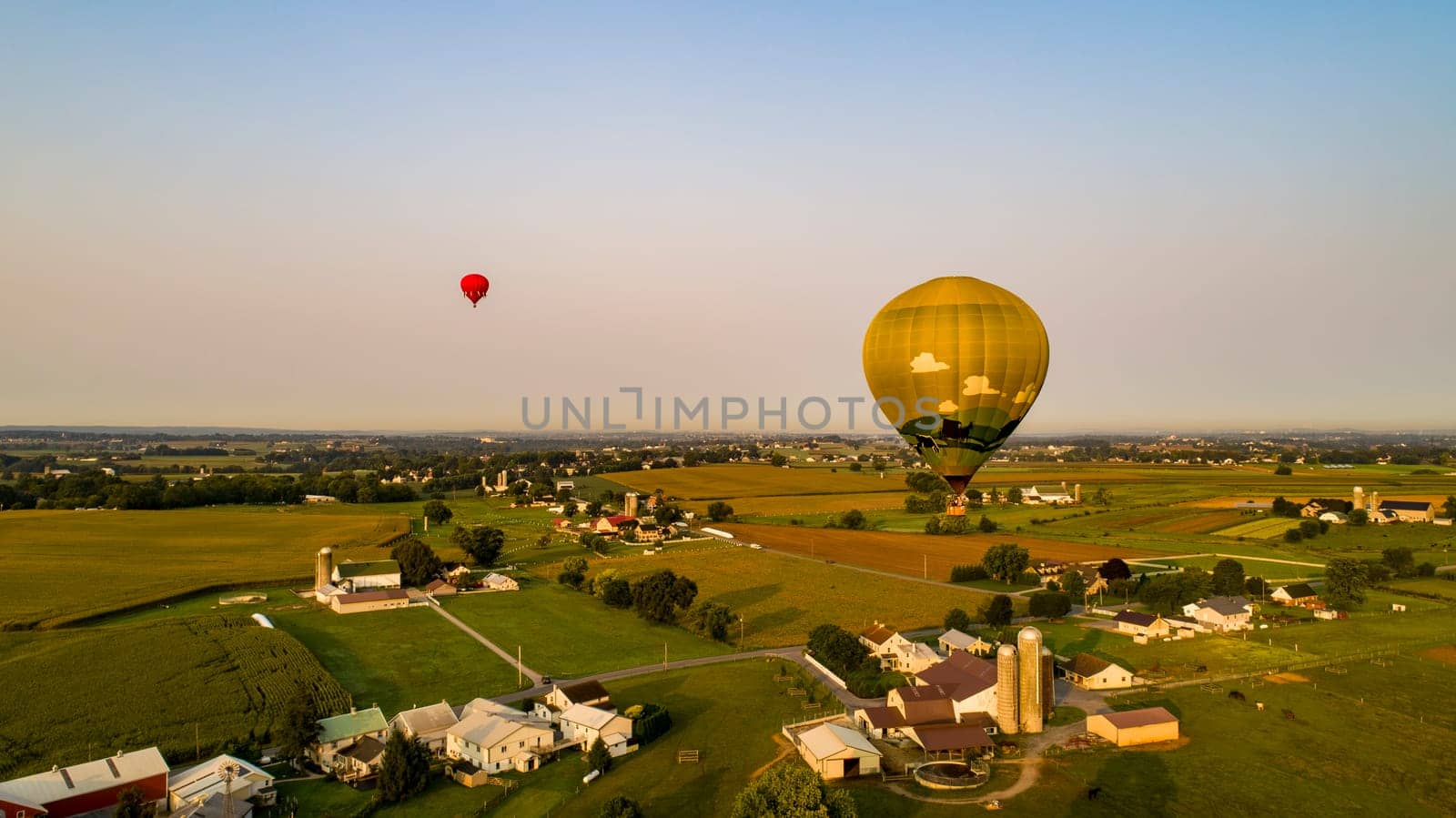 An Aerial View of Multiple Colorful Hot Air Balloons Floating Over Farm Fields of Corn and other Vegetables and Homes, on a Sunny Summer Morning