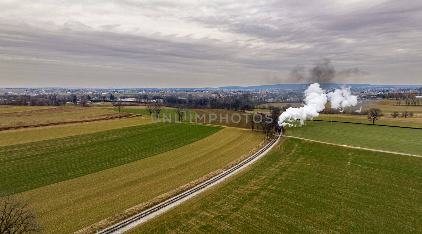 An Aerial View of a Steam Passenger Train Approaching, Traveling Thru Open Farmlands, Blowing Lots of White Smoke, on a Winter Day