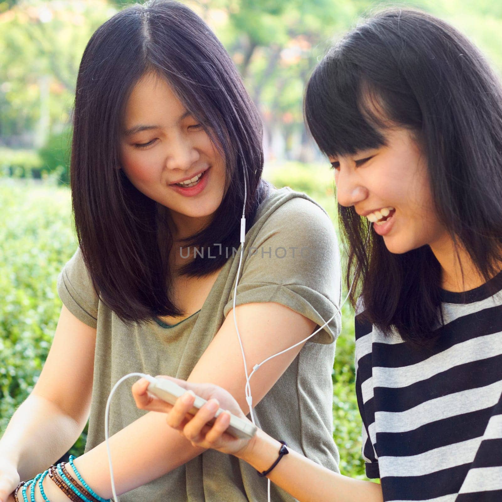 Asian women, students together and earphones with comic laugh, funny music video and meme at campus. Japanese girl, friends and audio streaming with smartphone, sharing and listen with smile in park.