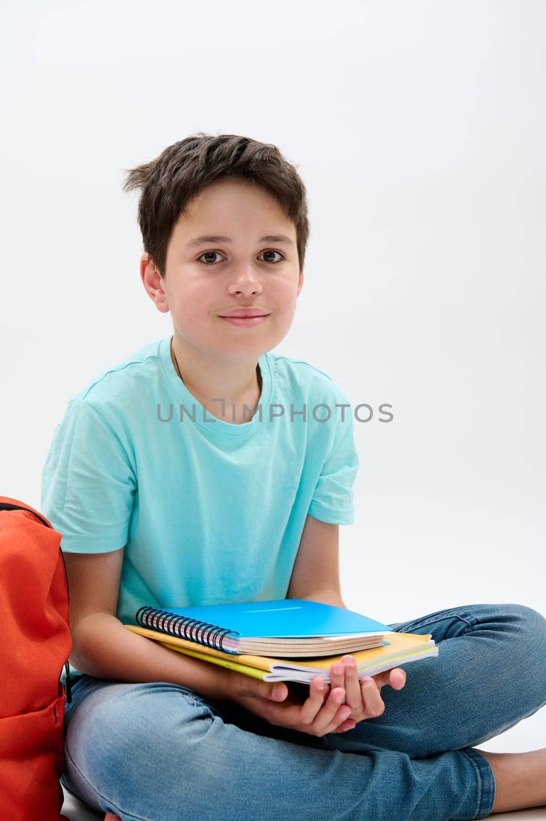 Vertical studio portrait of a handsome happy positive school kid boy holding school supplies, smiling looking at camera, isolated on white background. Back to school on new semester of academic year