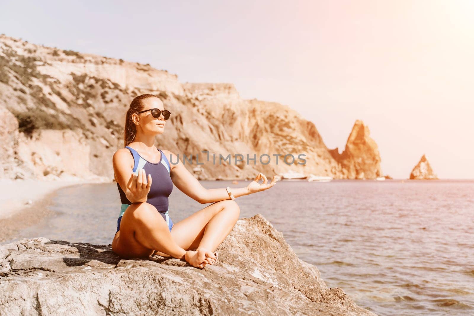 Yoga on the beach. A happy woman meditating in a yoga pose on the beach, surrounded by the ocean and rock mountains, promoting a healthy lifestyle outdoors in nature, and inspiring fitness concept