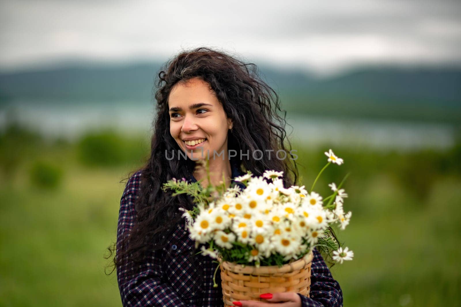 A woman stands on a green field and holds a basket with a large bouquet of daisies in her hands. In the background are mountains and a lake. by Matiunina