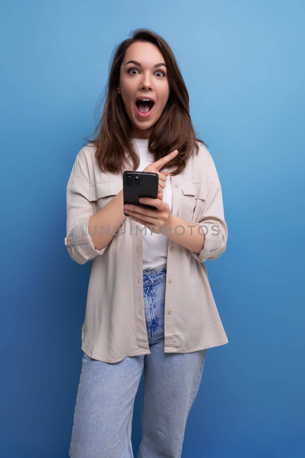 surprised caucasian young brunette woman in a shirt and jeans with a smartphone in her hands.