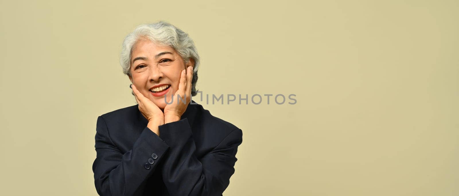 Smiling senior businesswoman wearing black suit and eyeglasses standing isolated on beige background.