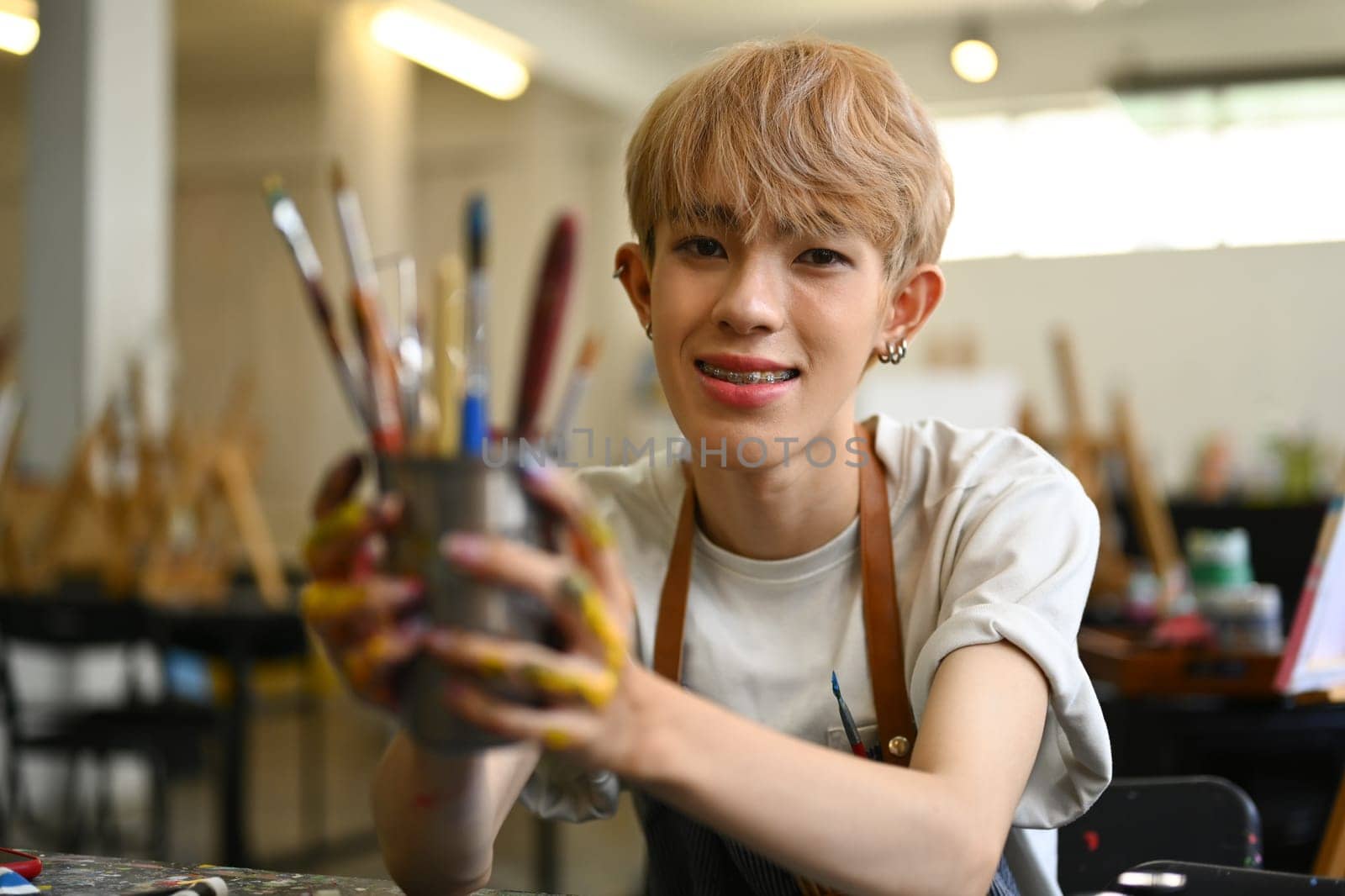 Pretty young gay man dressed in apron holding metal bucket with different brushes of the artist, selective focus.