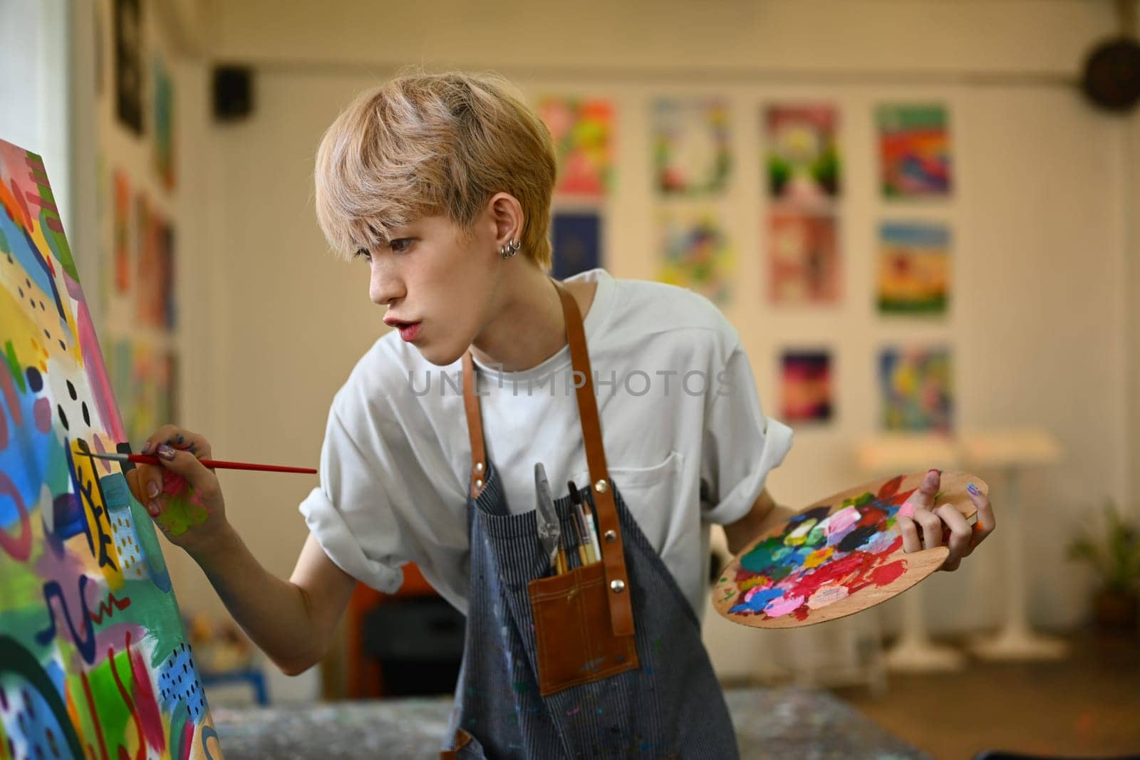 Concentrated teenage asian gay man dressed in apron painting picture with oil paints on canvas in bright art class.