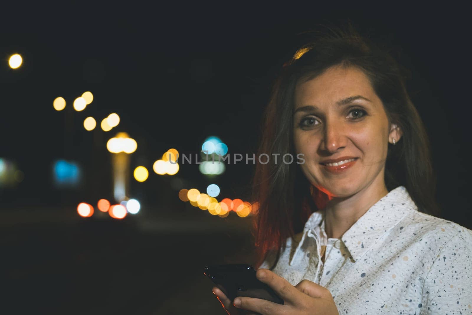 attractive girl in white suit with smartphone in night light reflection standing near copy space area for advertising, young woman smiling with mobile in hands