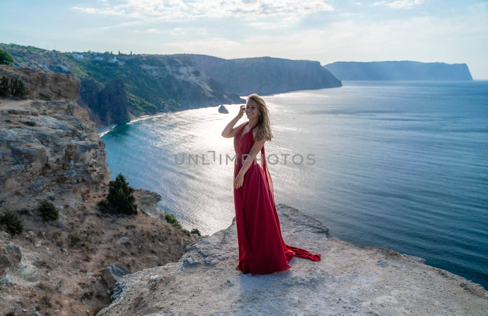 A girl with flowing hair in a long red dress stands on a rock above the sea. The stone can be seen in the sea. Sunny path to the sea from the sun. by Matiunina