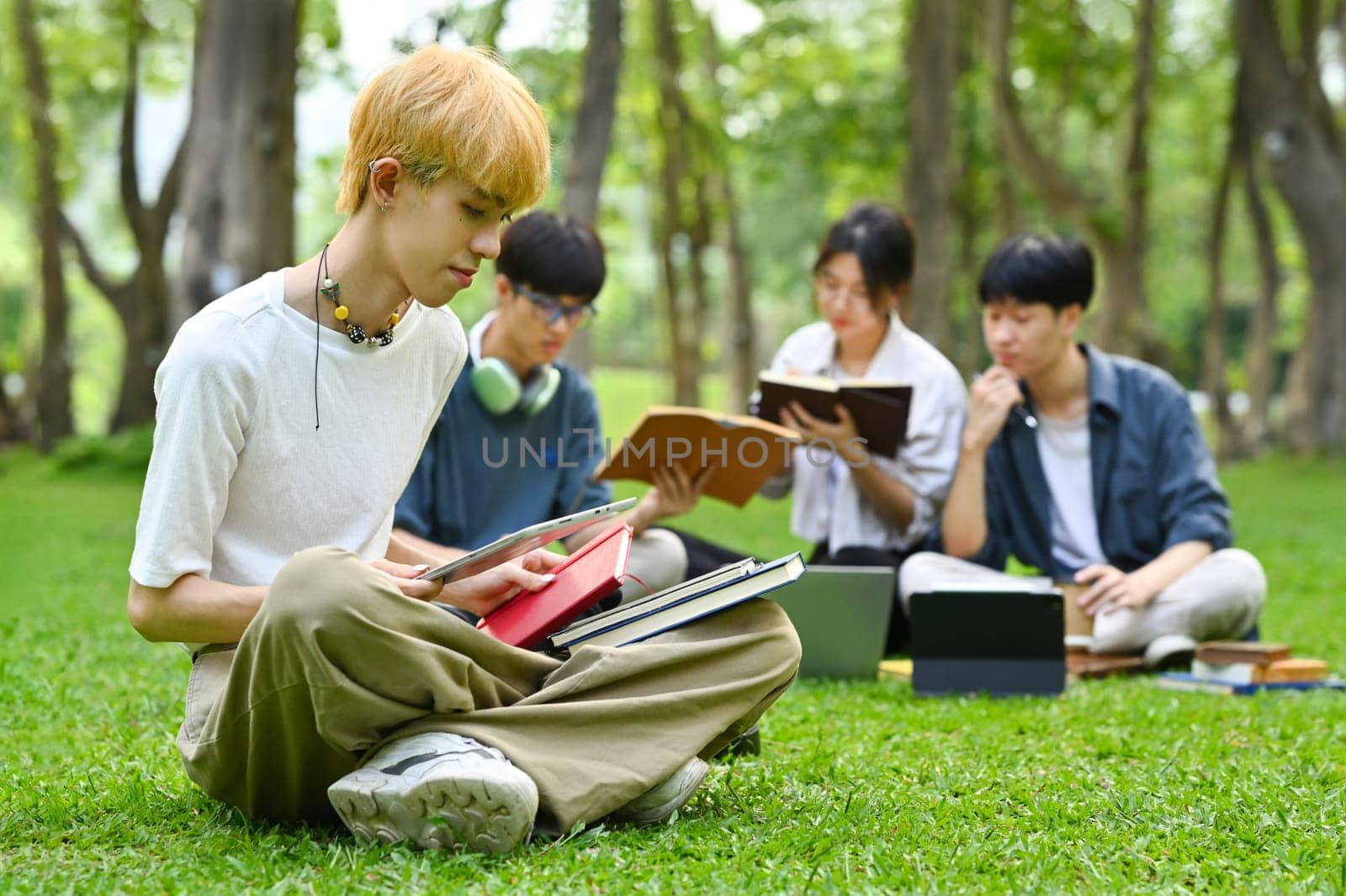 Asian male student sitting on green grass in campus with friends sitting on background. Education and lifestyle concept by prathanchorruangsak