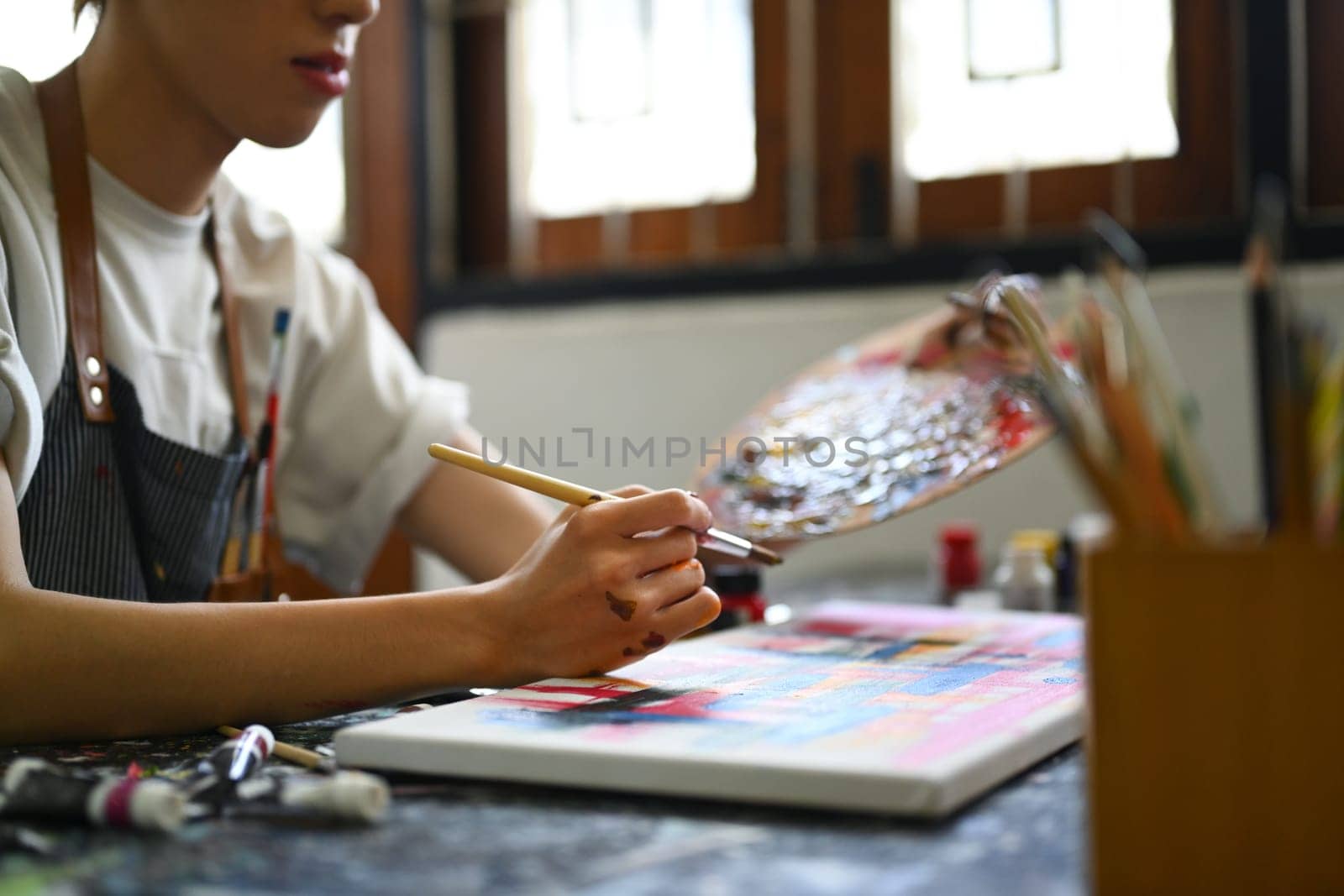 Cropped view of male artist holding messy dirty palette painting picture with brush on canvas at art studio by prathanchorruangsak