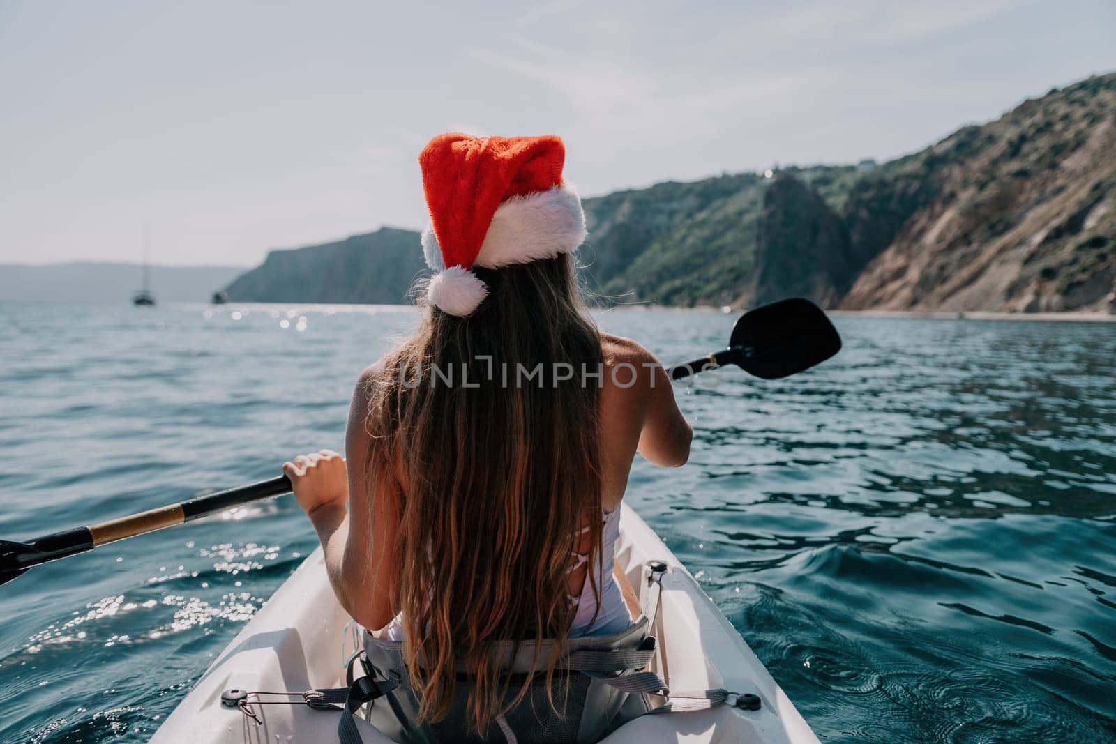 Woman in kayak back view. Happy young woman in Santa hat floating in kayak on calm sea. Summer holiday vacation and cheerful female people relaxing having fun on the boat. by panophotograph