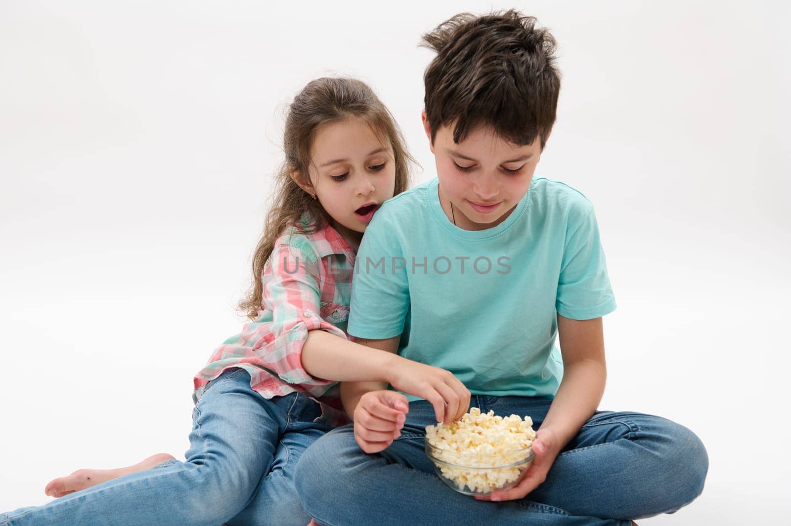 Beautiful kids, preteen boy and preschooler girl, dressed in casual clothes and blue denim jeans, eating tasty salted popcorn while watching movie, sitting together over isolated white studio backdrop