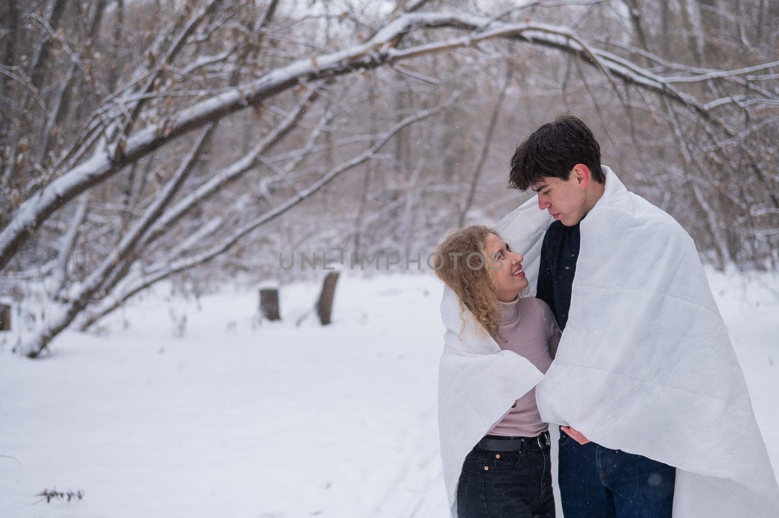 A young couple walks in the park in winter. The guy and the girl are kissing wrapped in a white blanket outdoors