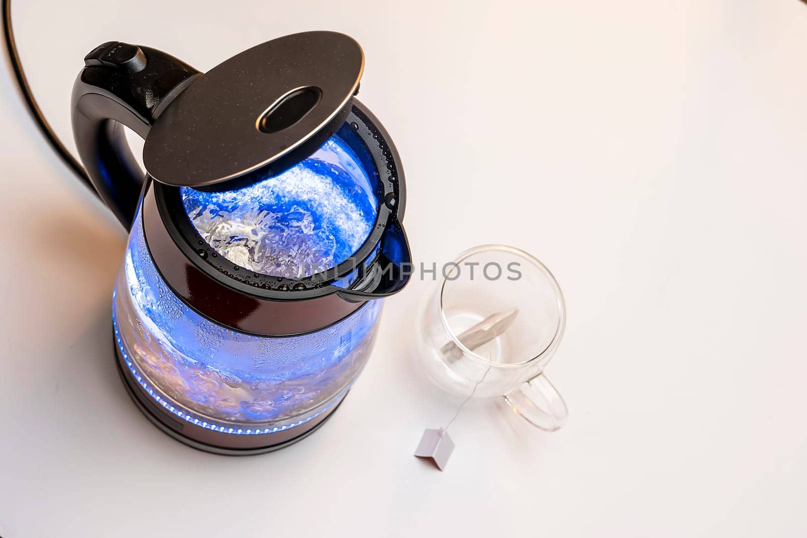 Brewing a tea bag. a fashionable tea cup with double glass walls and a modern electric transparent teapot with bubbling water and blue neon lighting on the table close-up.