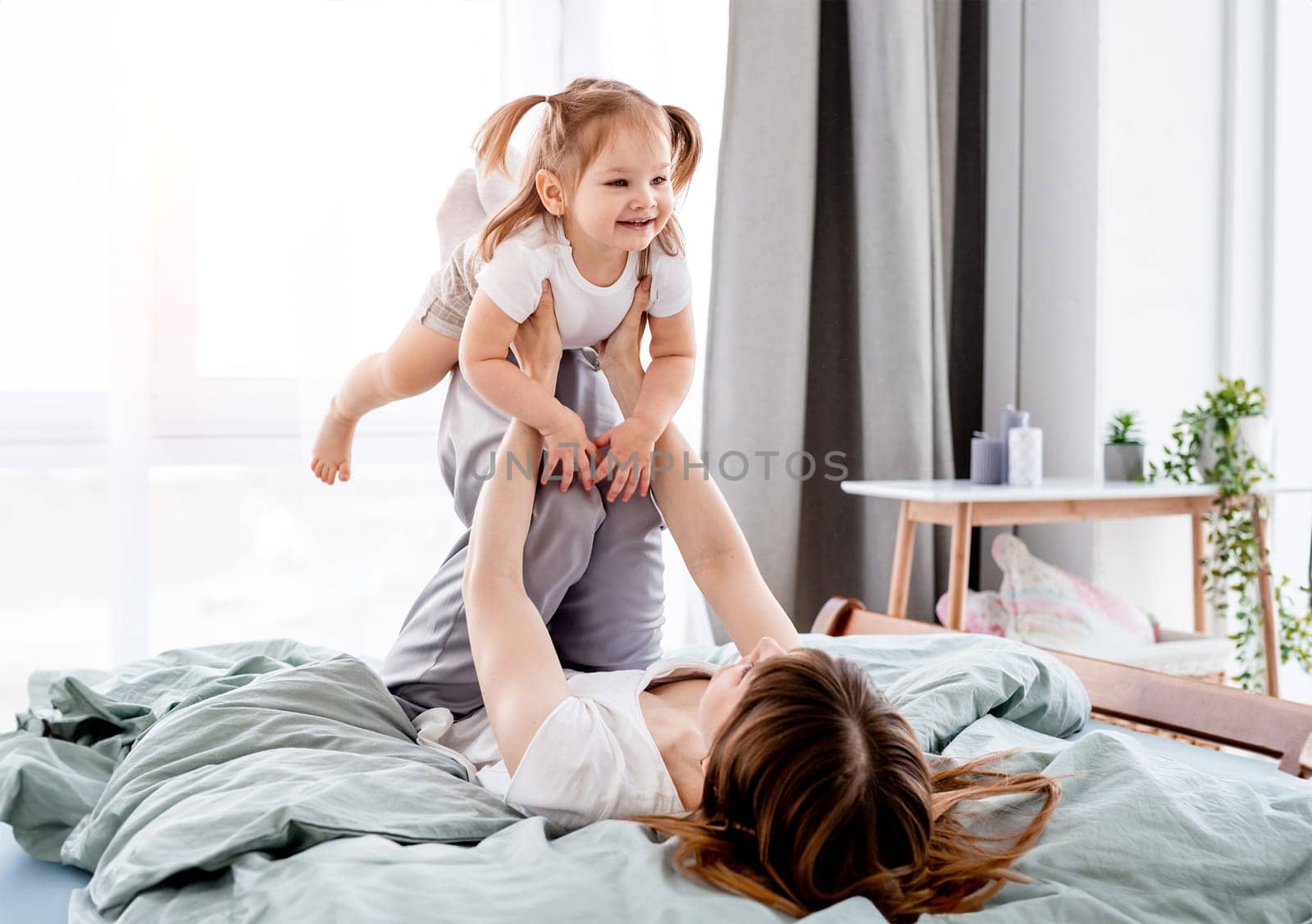 Young mother playing with daughter in the bed holding child on her legs. Mom and daughter spend time together in the bedroom with sunlight.