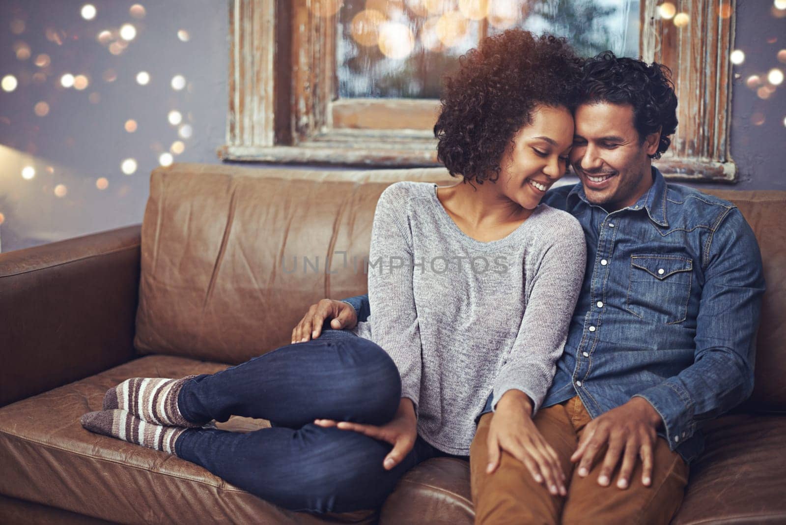 Hug, love and happy couple relax on a sofa, romantic and bonding on date night with bokeh. Interracial relationship, romance and man embrace woman on a couch, smile and chilling in a living room by YuriArcurs