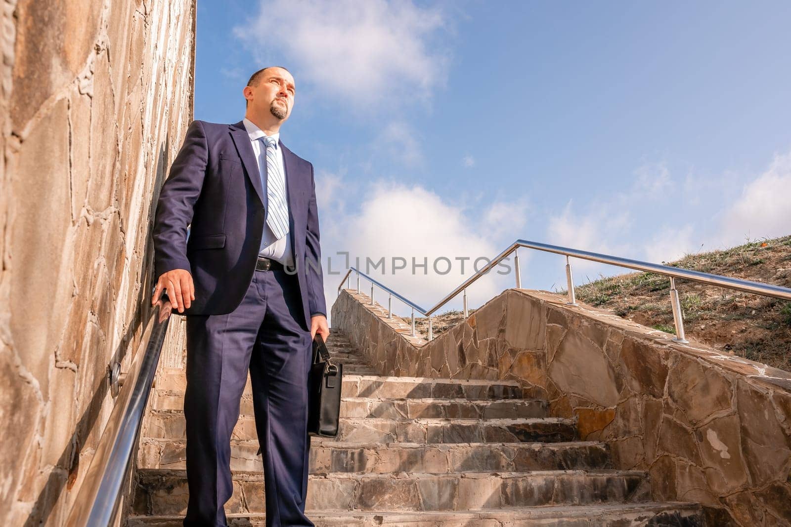 Confident middle age businessman with briefcase walking upstairs. Close-up of businessman wearing blue suit holding bag walking down the stairs by panophotograph