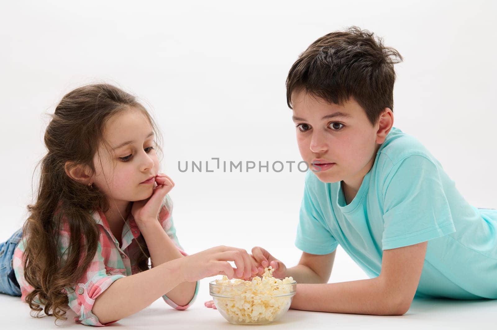 Beautiful happy children lying on belly, eating popcorn on isolated white background. Kids Childhood Lifestyle Leisure by artgf