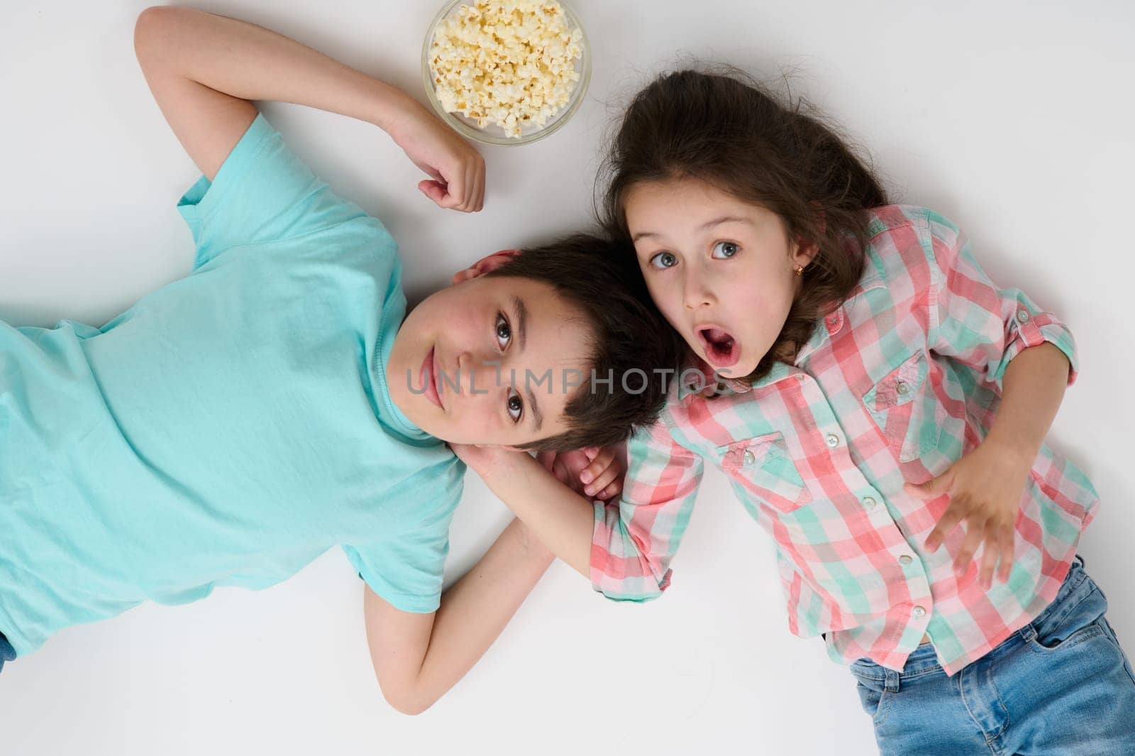 Happy children with popcorn bowl smile at camera, express surprise and amazement, lying on their backs on isolated white studio background. Kids. Childhood. Entertainment. Lifestyle. Leisure activity