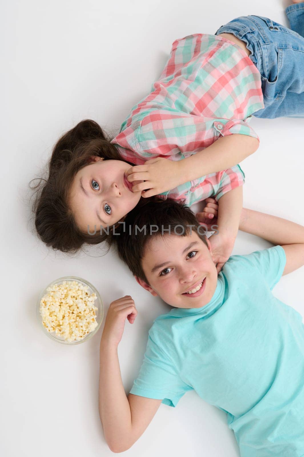 Top view happy kids, pre teen boy and little girl, brother and sister smiling looking at camera, lying on their backs on white studio background near a bowl with tasty popcorn, expressing positivity