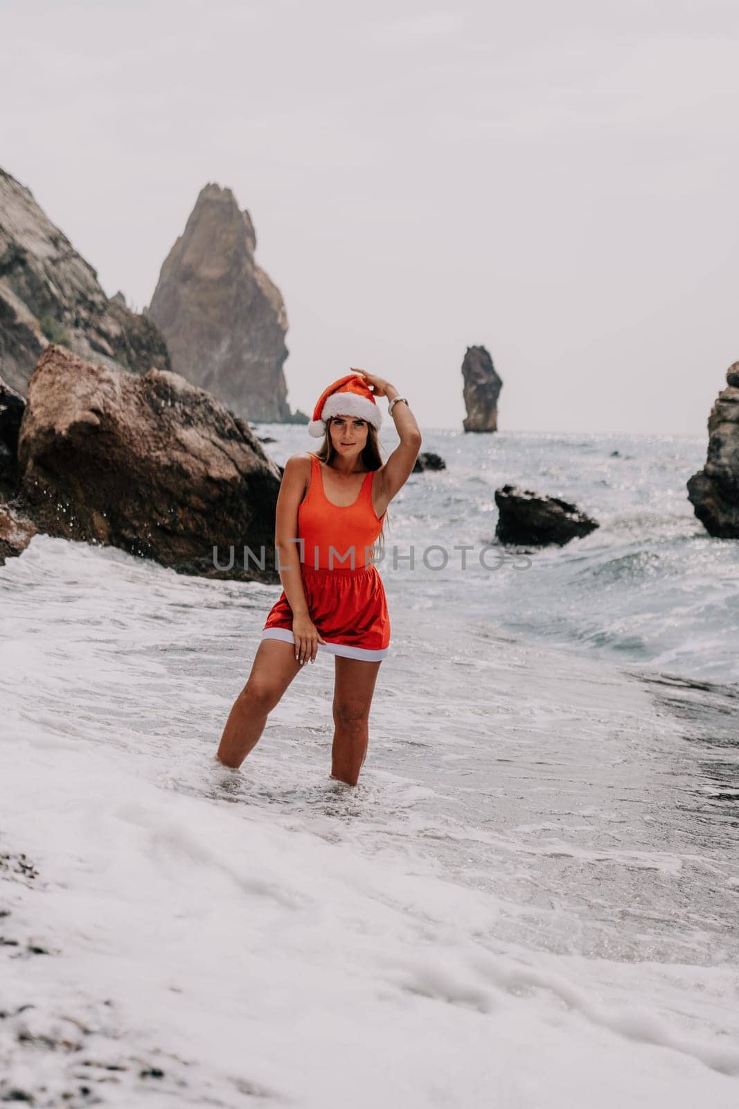 Woman summer travel sea. Happy tourist in red bikini and Santas hat enjoy taking picture outdoors for memories. Woman traveler posing on the beach surrounded by volcanic mountains, sharing travel joy by panophotograph