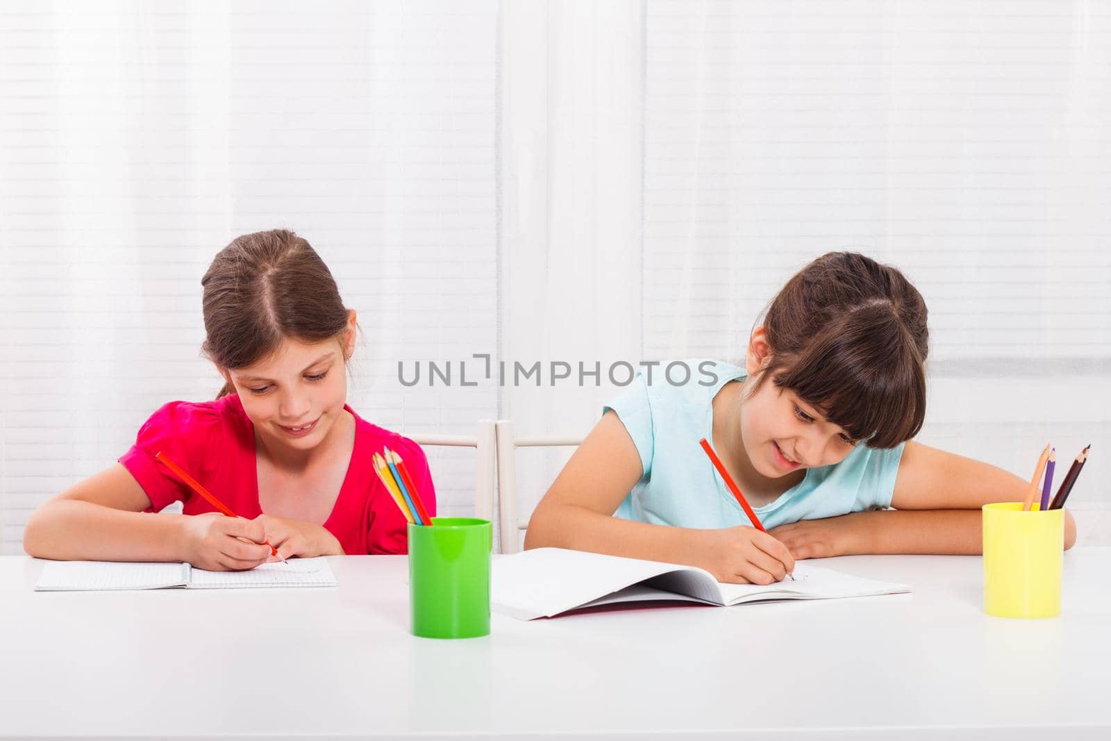 Two happy schoolgirls drawing at table in notebooks