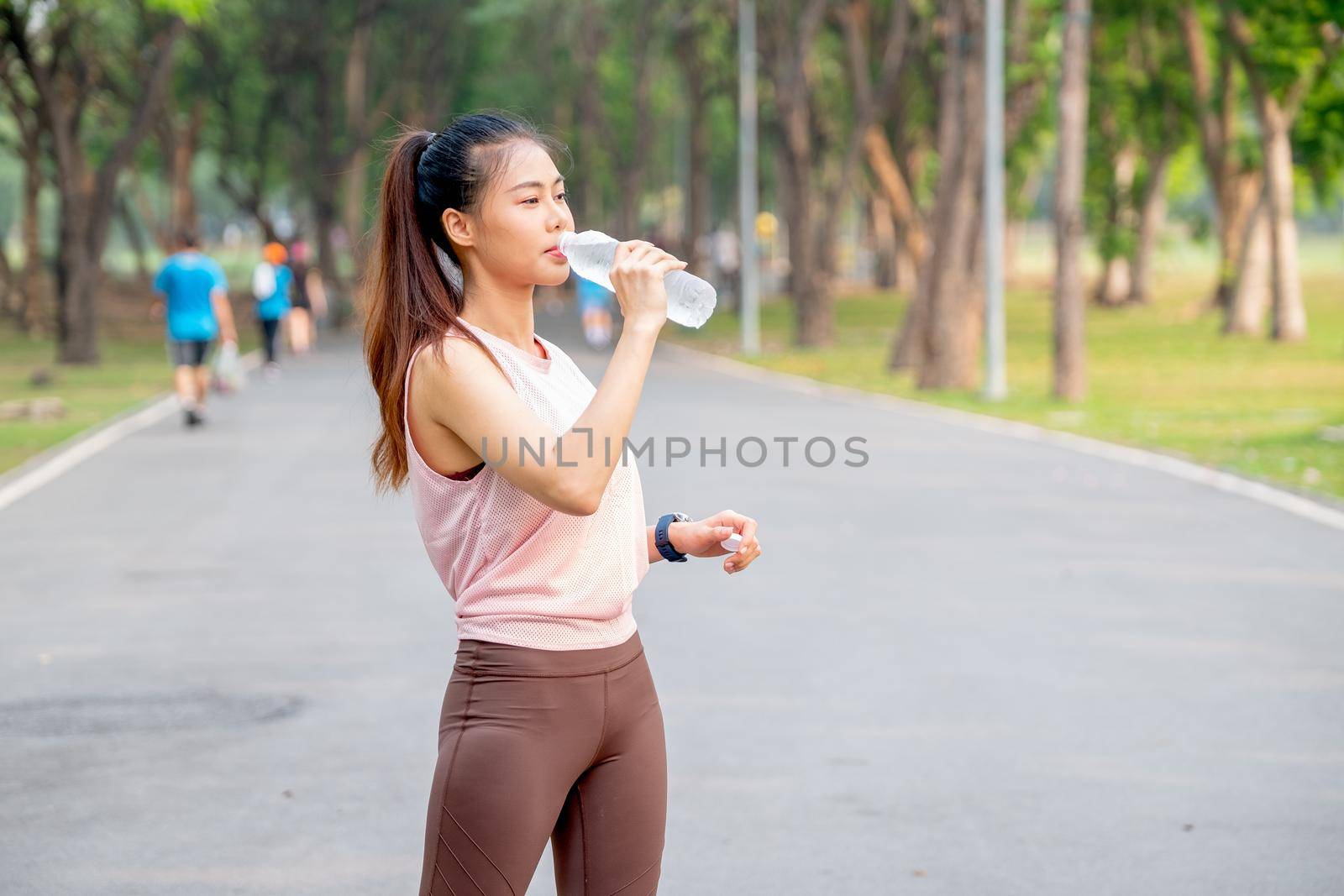 Asian woman with pink sportswear stand on road in park and drink water from bottle during exercise in the morning.