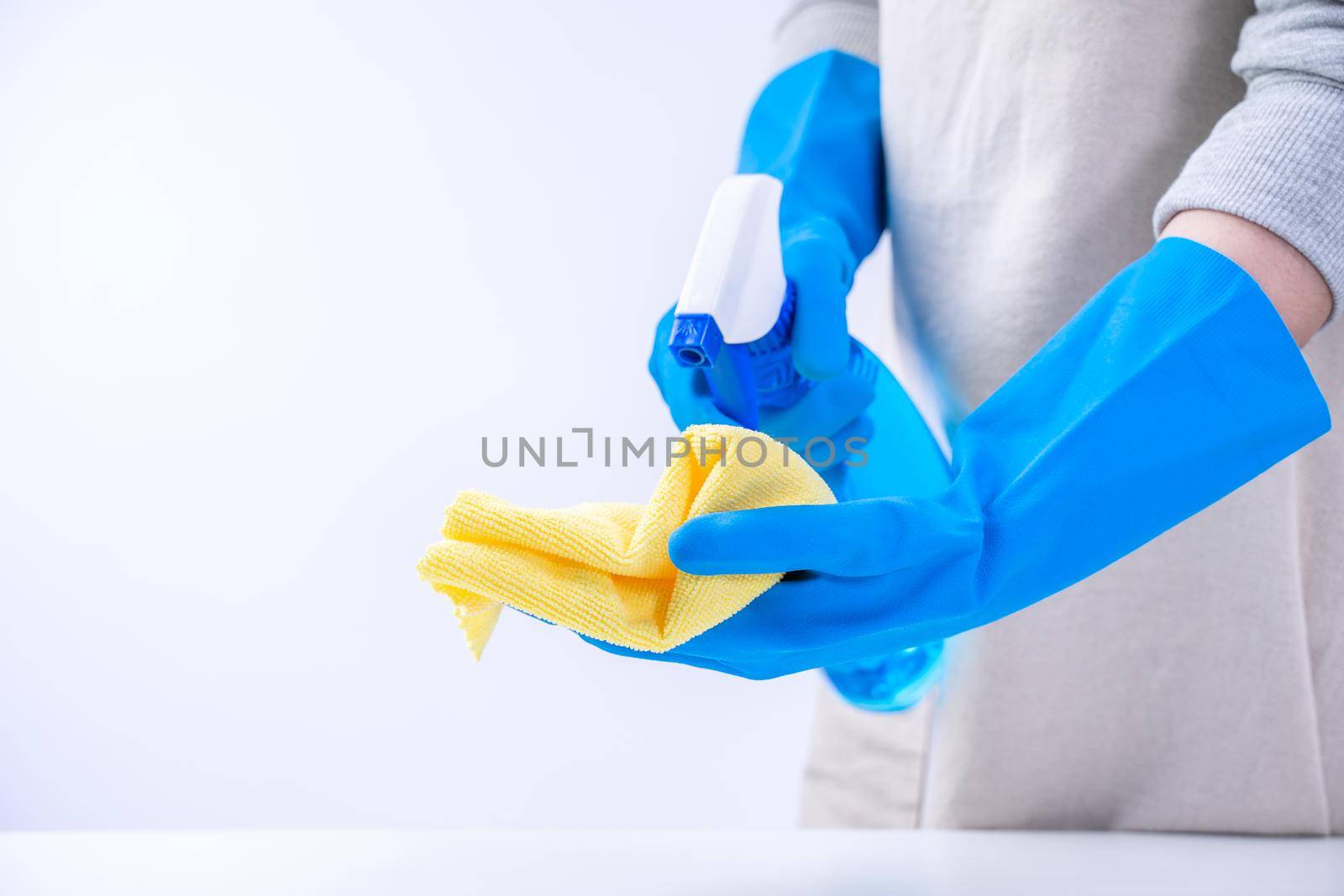 Young woman housekeeper is doing cleaning white table in apron with blue gloves, spray cleaner, wet yellow rag, close up, copy space, blank design concept. by ROMIXIMAGE