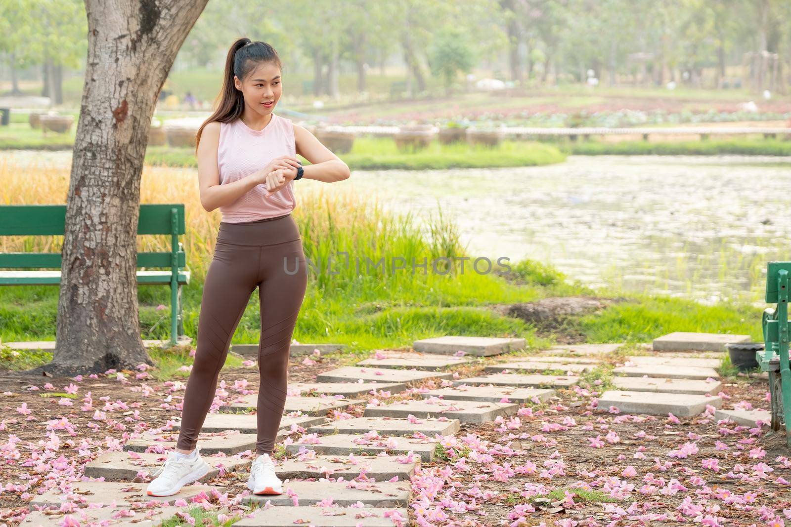 Wide shot of Asian woman stand and look at her watch during exercise in park or garden with green tree and lake as background in the morning.