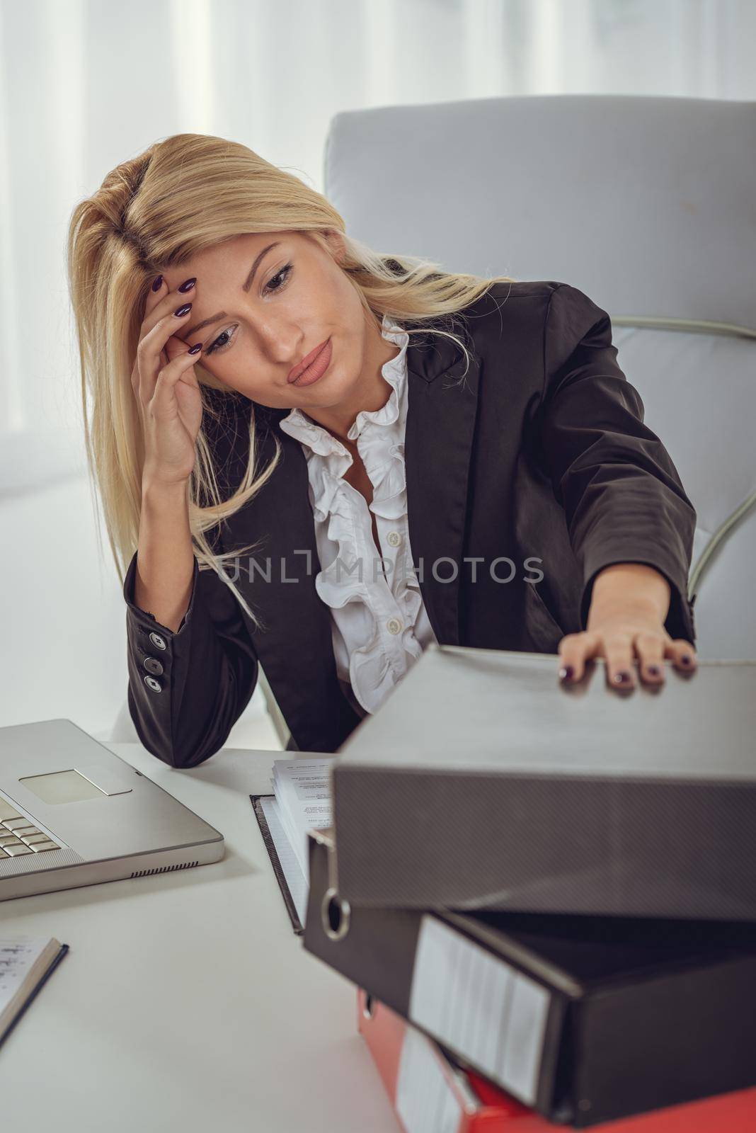 Young tired and worried business woman sitting in the office. There is a bunch of binders in front of her.
