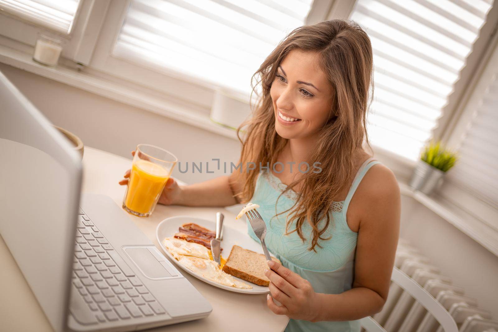 Young beautiful smiling woman having a breakfast at home and surfing social media at the laptop. On the plate there are bacon, eggs and slices of bread.