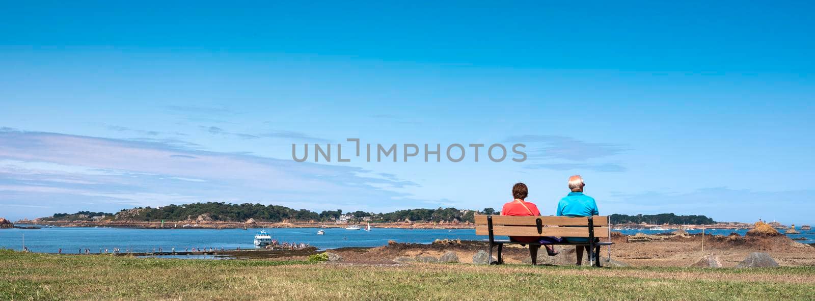 vedettes de bréhat, france, 10 august 2021: couple on bench looks at ferry to ile de brehat in french brittany under blue sky