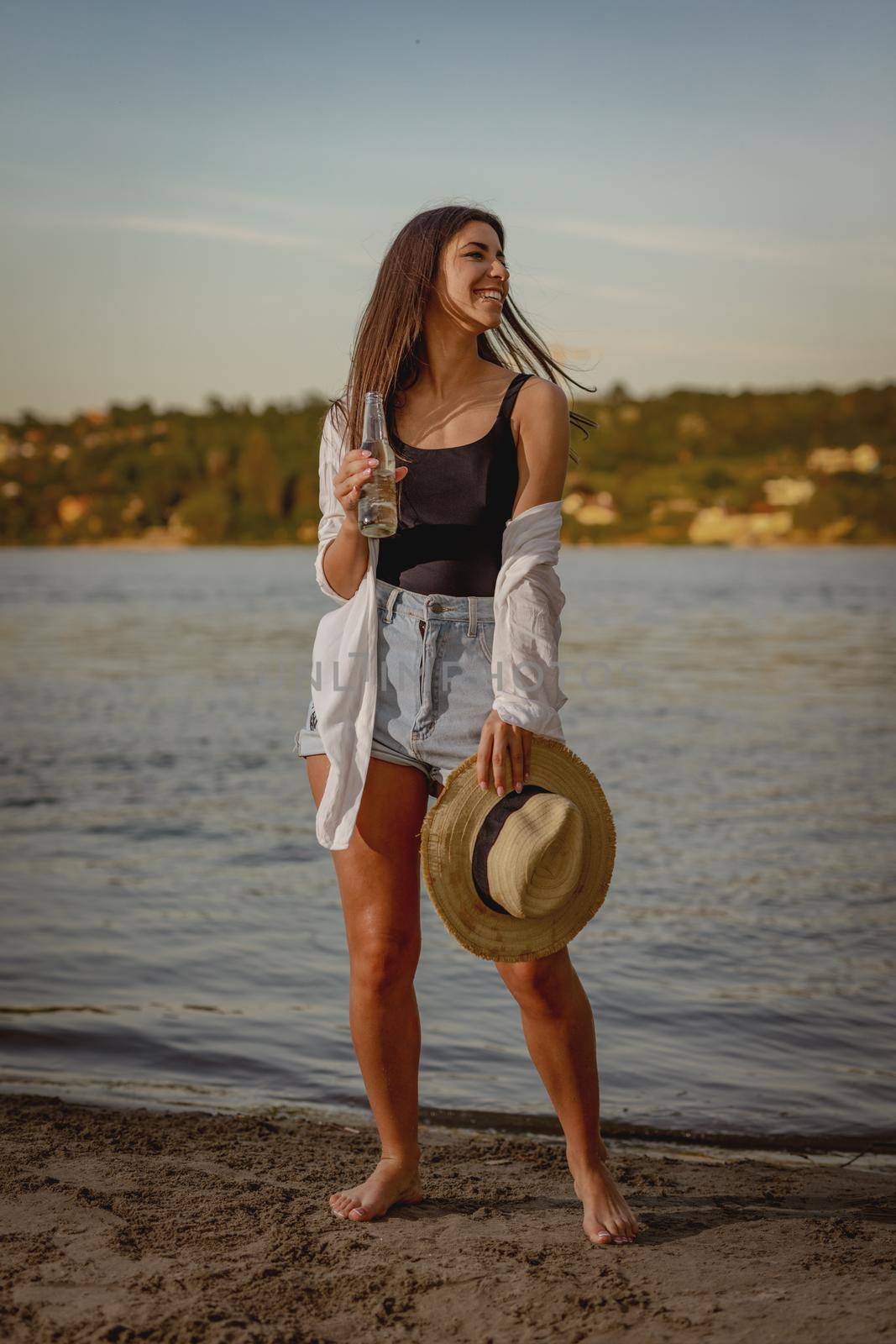 Portrait of a young smiling woman in straw hat relaxing at sunset time on the river bank.
