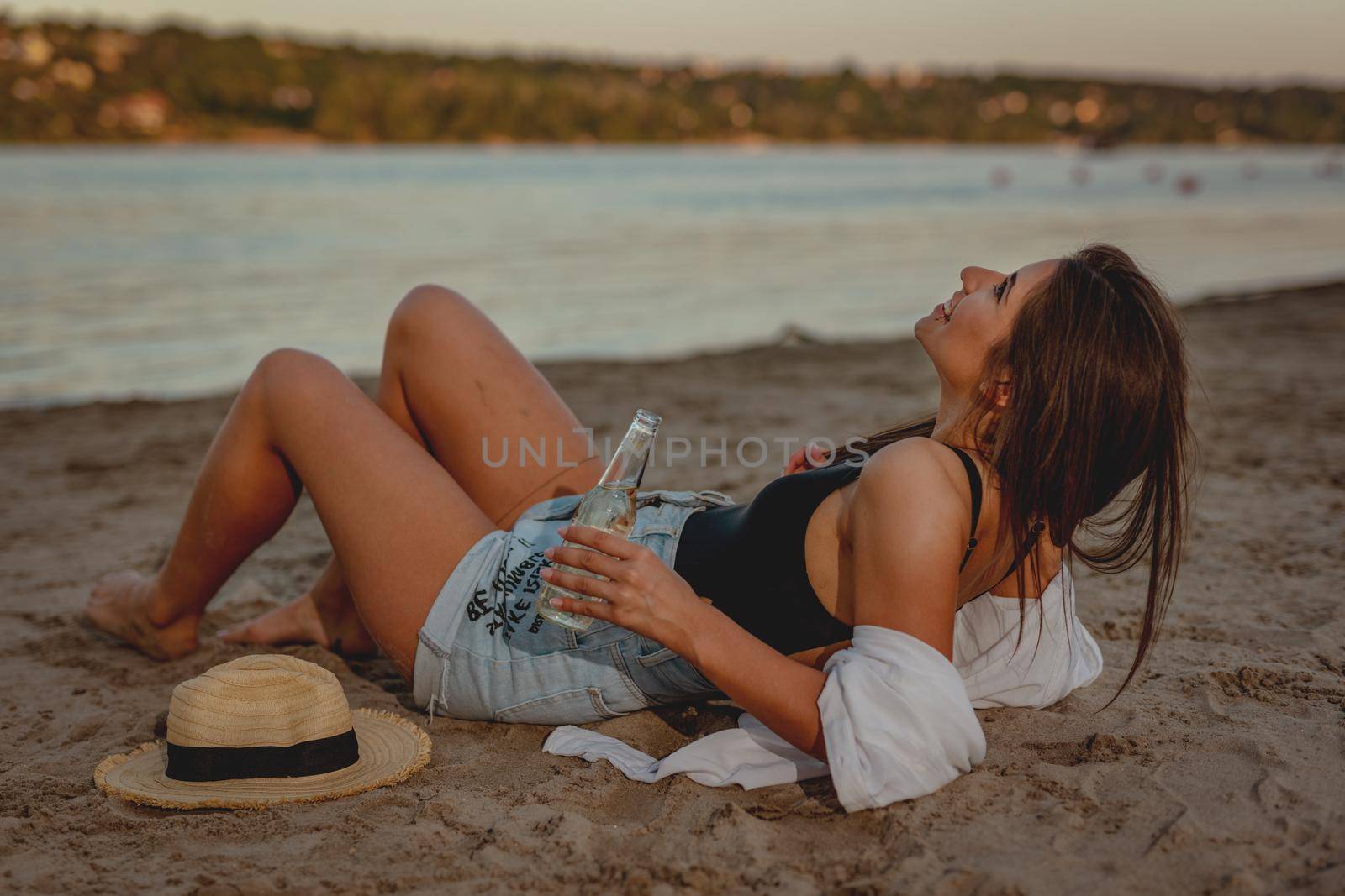 Young woman relaxing at sunset time on the river bank. She is lying down on the river beach and drinking beer. 