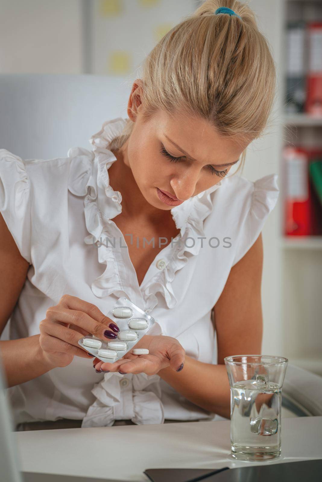 A young businesswoman with headache takes pills in her office, with a pained expression on her face.  