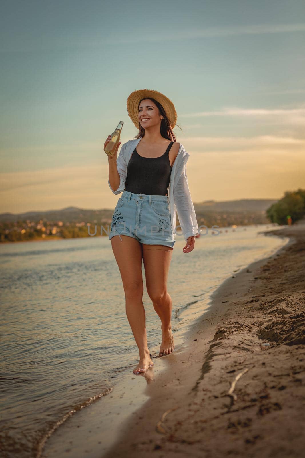 Young smiling woman in straw hat relaxing at sunset time on the river bank. She is walking along river beach and having fun.